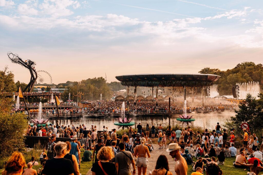 Multidão reunida no palco Crystal Garden durante o Tomorrowland Bélgica, com fontes, lago e decoração natural ao entardecer